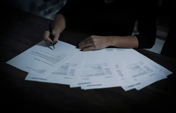 Hands signing through a fanned-out stack of corporate documents on a dark wood desk under low light