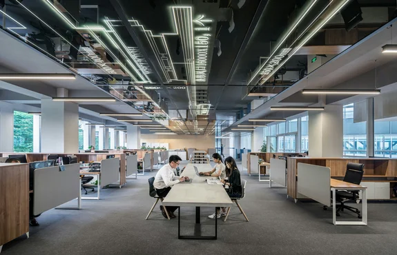 Two people working at a long table in a modern open-plan office with patterned overhead lighting
