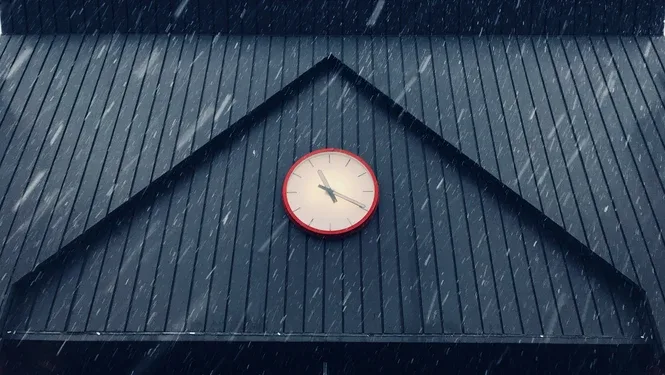 A red wall clock mounted on the dark gabled roof of a building with snow falling around it