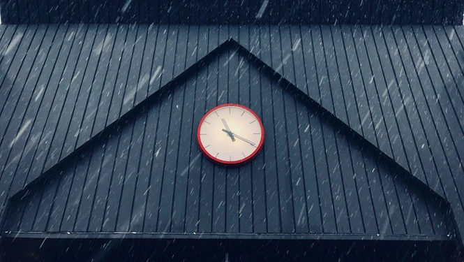 A red wall clock mounted on the dark gabled roof of a building with snow falling around it