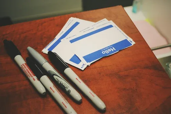 Black permanent markers beside a stack of blue and white 'Hello, my name is' name tags on a wood desk