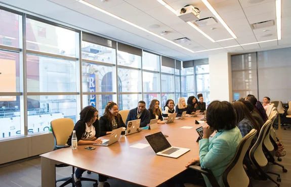 A diverse group meeting around a long conference table with laptops in a glass-walled boardroom