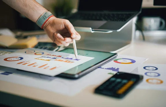 A hand holding a pen pointing at charts on a tablet, surrounded by printed graphs and a calculator