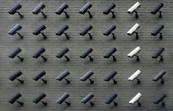A grid of dozens of black and white surveillance cameras mounted across a grey brick wall