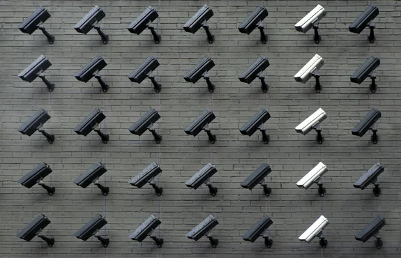 A grid of dozens of black and white surveillance cameras mounted across a grey brick wall