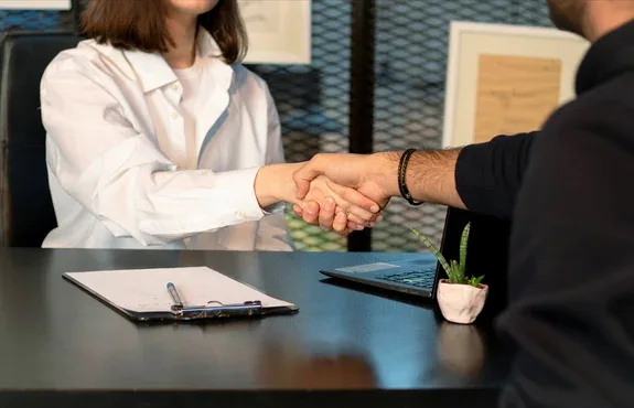 Two people shaking hands across a black office table over a clipboard and a small open laptop