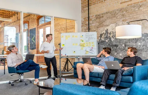 Four people gathered around a whiteboard covered in sticky notes in a loft-style office with exposed brick