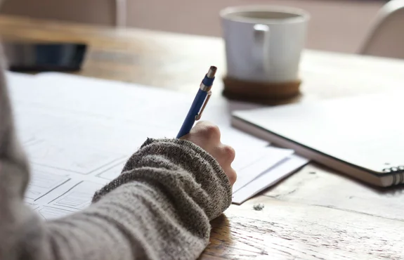 A hand in a grey knit sweater holding a pen over a printed document next to a white coffee mug