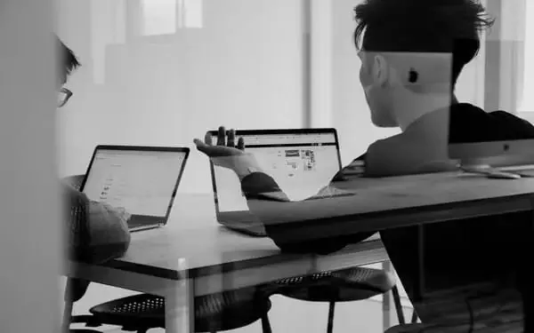Black-and-white view through a glass partition of two people working at laptops across a table